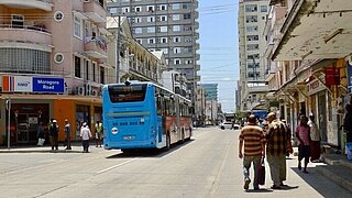 A busy city street with multi-story buildings, a blue public bus in the center of the road, and pedestrians walking along.