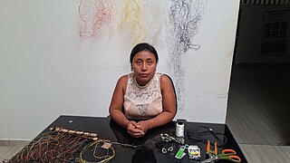 Woman sitting at a table with various tools and wire artworks in front of a white wall with hanging wire sculptures