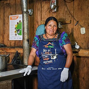 Frau mit buntem Kleid und Schürze steht vor einem Kochherd in einer Holzhütte in Guatemala