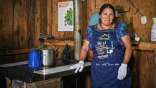 Woman wearing colorful dress and apron standing in front of a cookstove inside a wooden hut in Guatemala