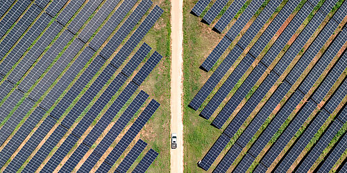 An aerial view of a solar farm, with rows of solar panels placed on the land and a car on the road crossing the middle.