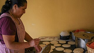 A woman with a braid and checkered apron baking tortillas on a countertop with multiple tortillas in a kitchen in Guatemala.