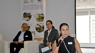 Drei Personen sitzen bei einer Veranstaltung, eine Frau spricht in ein Mikrofon, im Hintergrund ein Banner mit Text und Bildern zu landwirtschaftlichen Themen