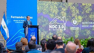 Ein Sprecher auf einer Bühne steht am Pult und stellt den Fortschritt der NDC 3.0 in El Salvador im Mai 2025 vor. 
