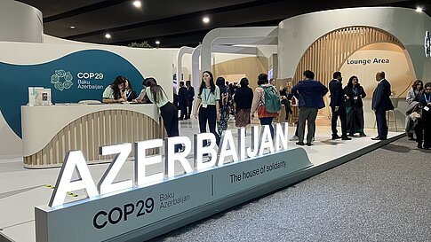 Eine große Messestandfläche bei der COP29 mit dem Schriftzug „AZERBAIJAN“ in leuchtenden Buchstaben im Vordergrund. Dahinter der offizielle COP29-Stand mit mehreren Personen, die sich unterhalten oder an einem Empfangstresen arbeiten. Auf der Wand steht „COP29 Baku Azerbaijan“ und „The house of solidarity“. Im Hintergrund befinden sich weitere Besucher*innen im Gespräch und eine Lounge Area. Die Szenerie ist lebendig und geschäftig.