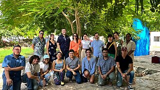 Eine Gruppe von siebzehn Personen posiert im Freien unter einem großen, belaubten Baum, durch dessen Zweige Sonnenlicht fällt. Acht Personen knien oder sitzen in der vorderen Reihe, während neun dahinter stehen und freundlich in die Kamera lächeln. Die Gruppe ist leger gekleidet, und die Umgebung wirkt entspannt und sonnig mit viel Grün und einer leuchtend blauen Wand im Hintergrund.