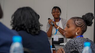 Eine Frau mit Mikrofon trägt in einem Konferenzraum etwas vor und das Publikum hört zu.