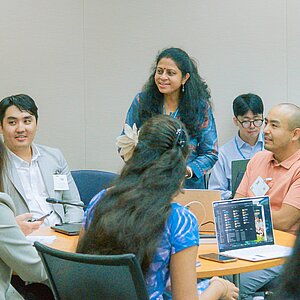 Teilnehmende eines Workshops unterhalten sich in einer Kleingruppe; einige machen Notizen, andere sehen auf Laptops, in einer freundlichen, konzentrierten Atmosphäre.