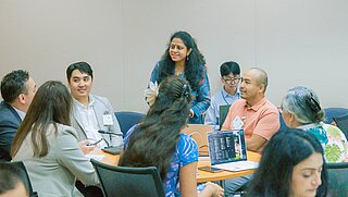 Teilnehmende eines Workshops unterhalten sich in einer Kleingruppe; einige machen Notizen, andere sehen auf Laptops, in einer freundlichen, konzentrierten Atmosphäre.