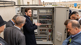 A group of people gathers around a refrigeration system, while a man in a dark suit points to and explains the various components and cables inside.