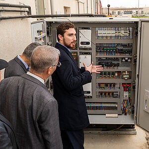 A group of people gathers around a refrigeration system, while a man in a dark suit points to and explains the various components and cables inside.