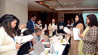 Mehrere junge Frauen und Männer gruppieren sich in einem Konferenzraum um einen Tisch herum und führen Verhandlungen.