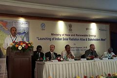 Person am Rednerpult, daneben sitzen fünf Personen am Tisch