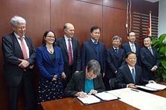 Hendricks und Zhenggao sitzen am Tisch, um sie herum steht Gruppe von Menschen