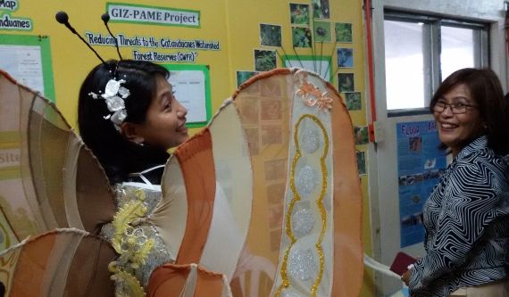 Zwei lachende Frauen vor einem Projektplakat. Eine trägt große bunte Schmetterlingsflügel