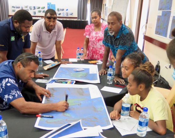Studierende und andere Menschen aus Vanuatu stehen und sitzen um einen Tisch mit Kartenmaterial und arbeiten hieran gemeinsam.