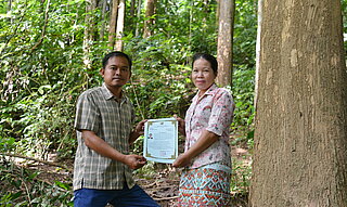 Ein Mann und eine Frau stehen neben einem Baum und halten ein Zertifikat in der Hand.