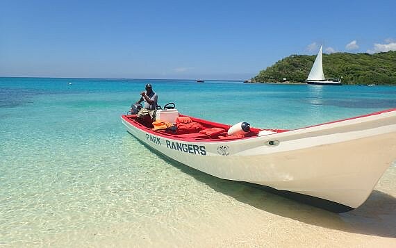 Ein offenes Boot mit der Aufschrift Park Ranger steht am Strand in seichtem türkisfarbenen Wasser des karibischen Meeres, ein Mann sitzt darin, im Hintergrund verläuft eine bewaldete Landzunge ins Meer, vor der ein Segelboot mit gehisstem Vorsegel fährt. Die Park Ranger sind auf die Mitwirkung der Bevölkerung angewiesen, um Küsten und Strände sauber zu halten. St. Vincent und die Grenadinen. Foto: The Nature Conservancy.